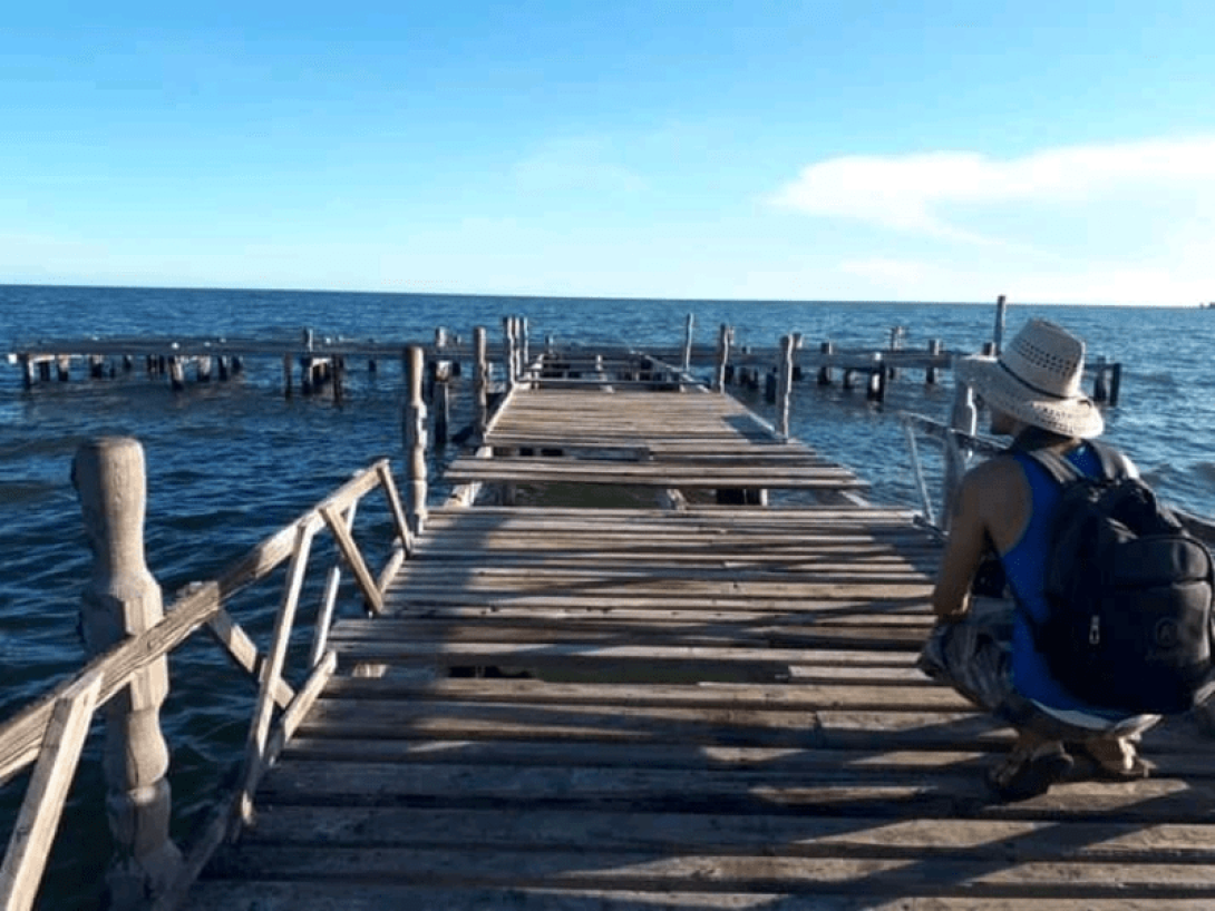 Persona agachada de espaldas en un muelle de madera frente al mar.