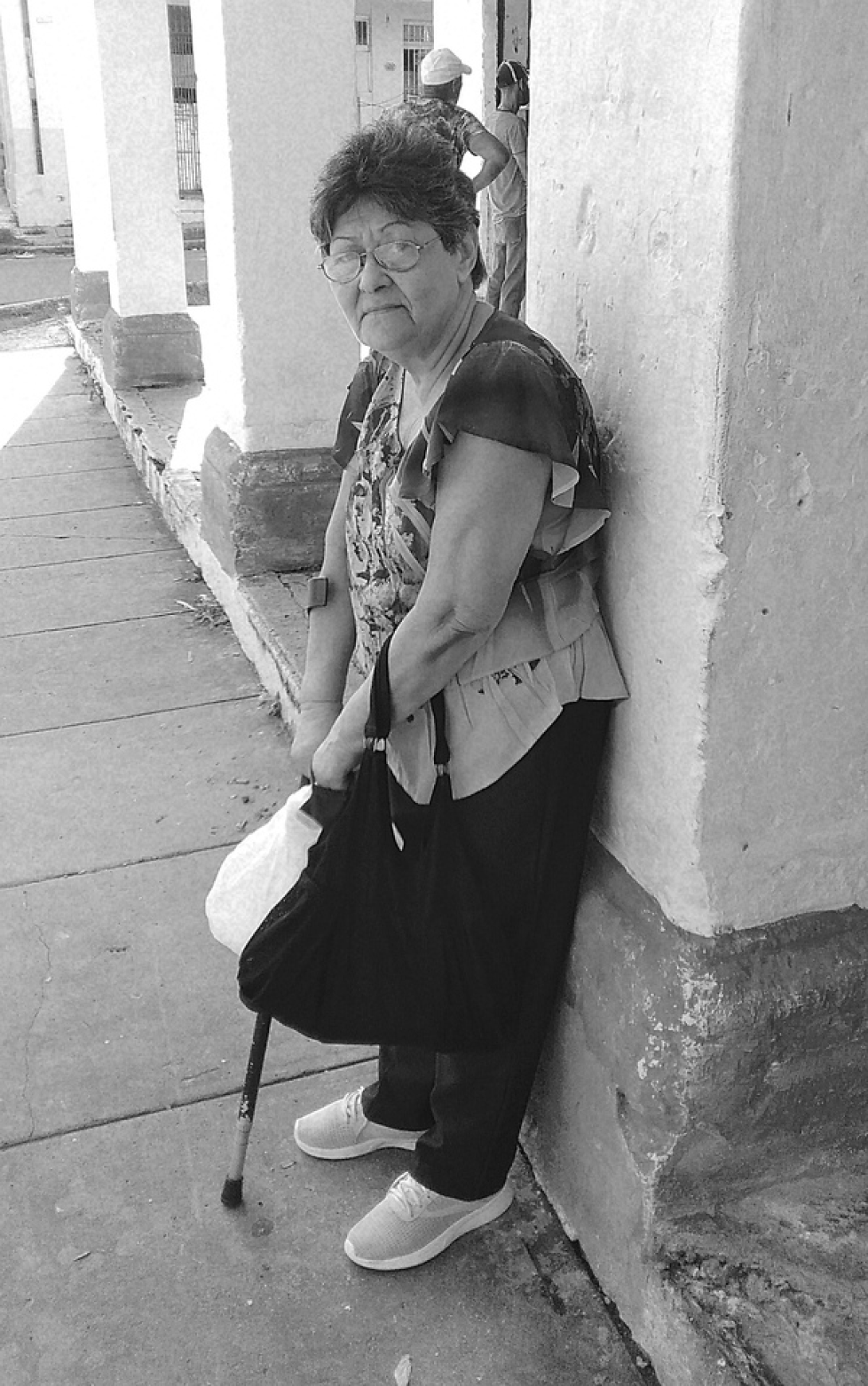 Anciana en Cuba, con bastón y bolsa de la compra, está recostada a una columna con cara de cansancio.