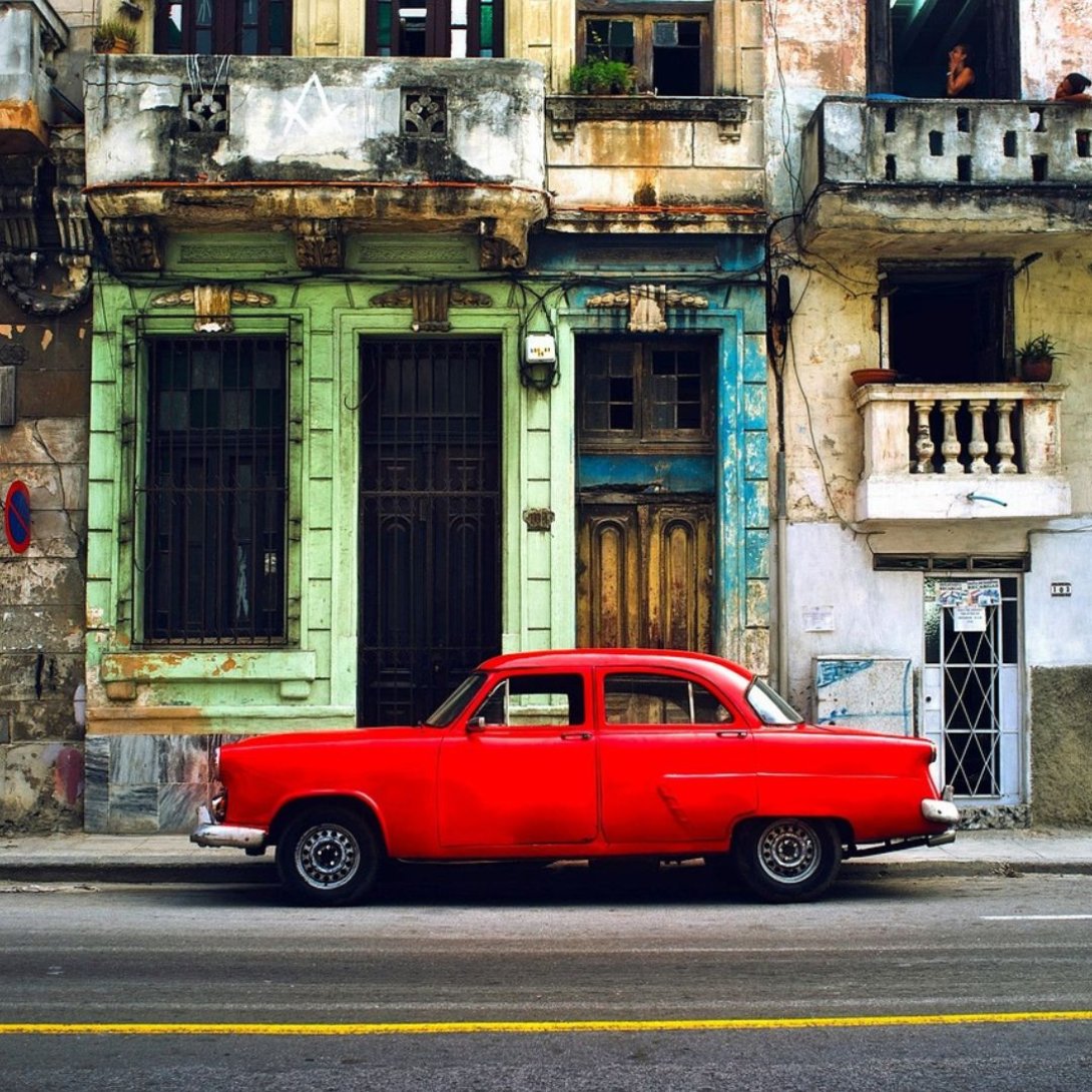 Automóvil rojo antiguo estacionado frente a fachadas desgastadas en una calle de Cuba, símbolo de la preservación y adaptación mecánica en el país.
