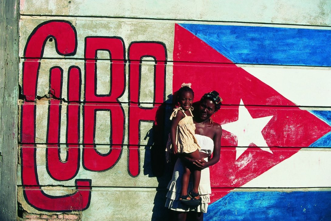 Mujer cargando una niña frente a una pared con la bandera cubana