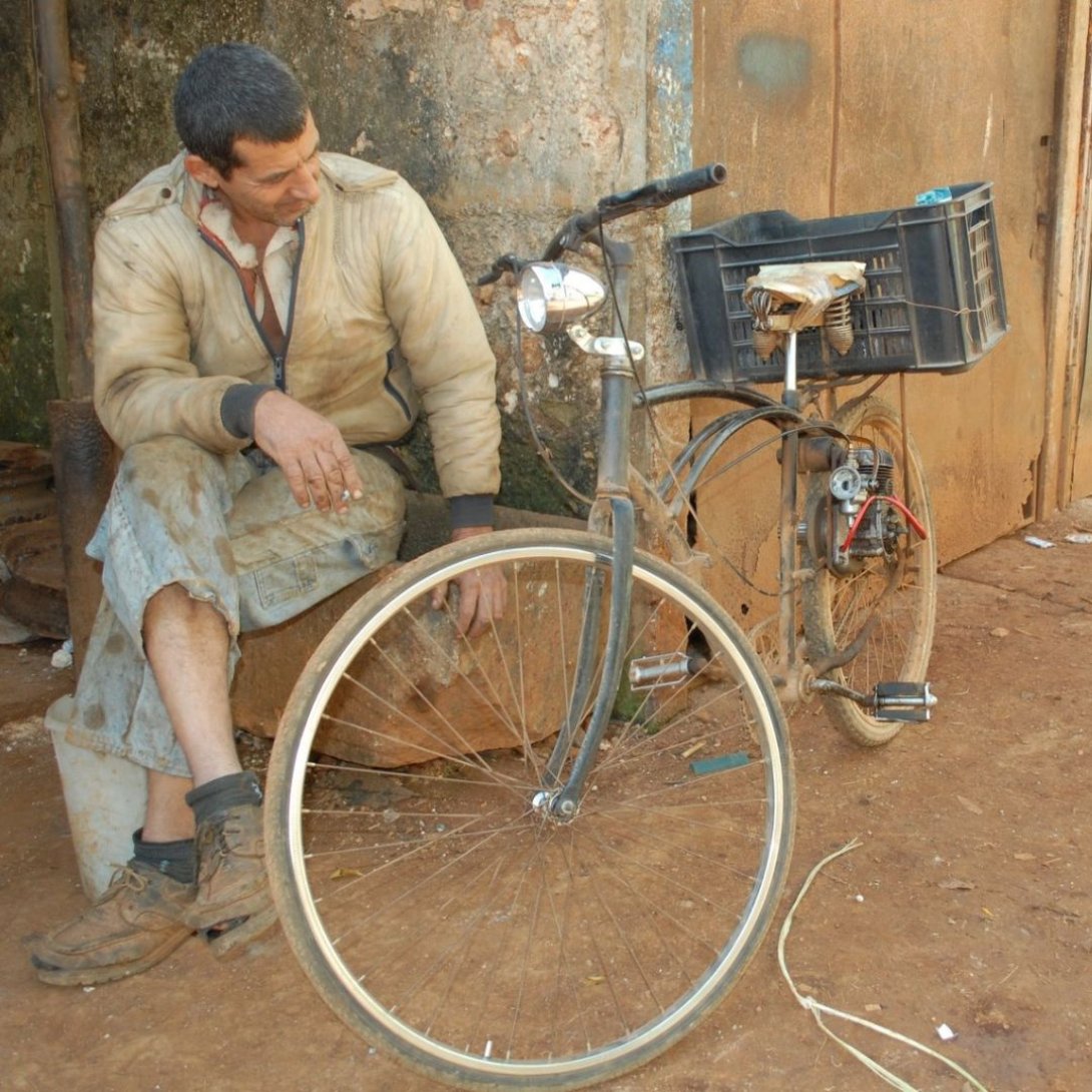 Hombre sentado junto a una bicicleta modificada artesanalmente con un motor y una caja de plástico, conocida como rikimbili.