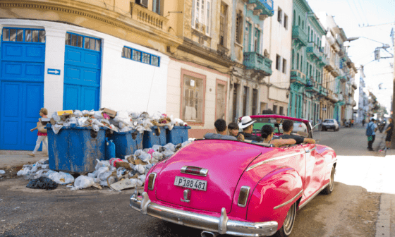 Carro con turistas pasa por un basurero en una esquina de una calle.