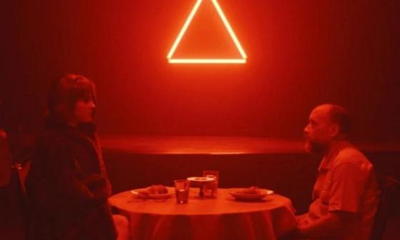 Un hombre y una mujer conversan de frente sentados en una mesa de una habitación con luz roja. 