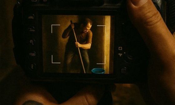 Imagen en perspectiva: un hombre trapeando visto a través del visor de una cámara réflex, sostenida por una mano tatuada con la Virgen.