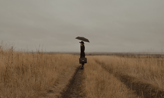 Una mujer con sombrilla y maleta en mano camina por un campo. 