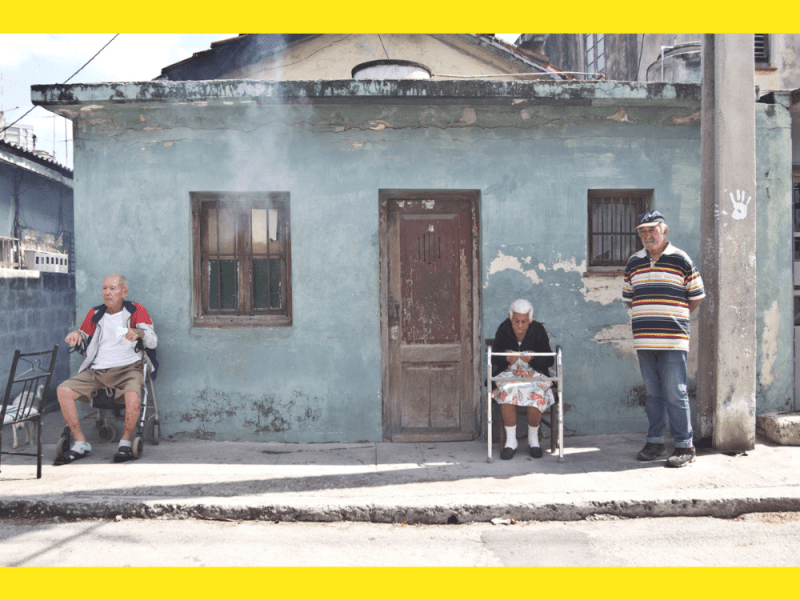 Ancianos esperan sentados en la calle mientras fumigan sus casas, Cuba. Sale humo por la ventana de una casa.